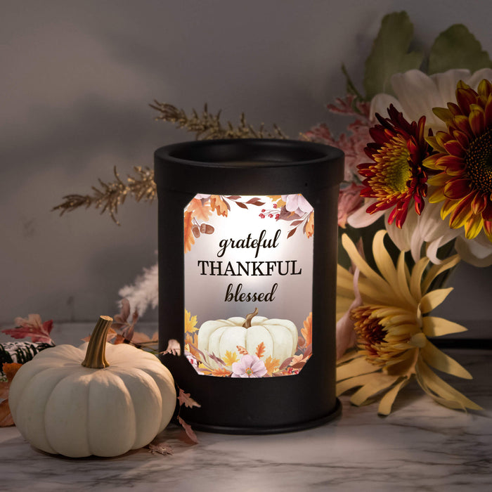A black cylindrical wax warmer with a  label that reads 'grateful THANKFUL blessed' and features a pumpkin illustration, styled on a marble surface with a small white pumpkin and autumn flowers.