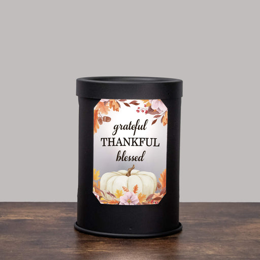 A black cylindrical wax warmer lamp standing on a wooden surface against a light gray background, with a decorative front label that reads 'grateful THANKFUL blessed' around a white pumpkin and autumn leaves.