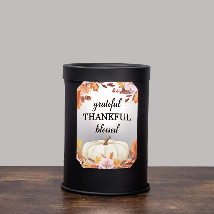 A black cylindrical wax warmer lamp standing on a wooden surface against a light gray background, with a decorative front label that reads 'grateful THANKFUL blessed' around a white pumpkin and autumn leaves.