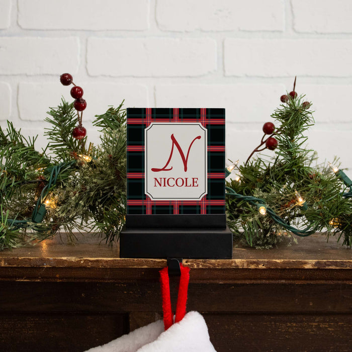 A personalized Christmas-themed stocking hanger hook with a black and red plaid pattern, featuring a white central panel with a red monogram 'N' and the name 'NICOLE'. The sign is displayed on a wooden mantel decorated with green pine branches, red berries, and warm string lights, with a white stocking hanging below.