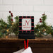 A personalized Christmas-themed stocking hanger hook with a black and red plaid pattern, featuring a white central panel with a red monogram 'N' and the name 'NICOLE'. The sign is displayed on a wooden mantel decorated with green pine branches, red berries, and warm string lights, with a white stocking hanging below.