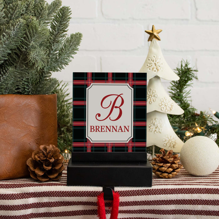 A personalized Christmas-themed stocking hanger hook, featuring a red and green plaid border with a white center panel and the monogram 'B' and name 'BRENNAN' in red. The item is styled on a holiday mantel with pine branches, a white decorative tree, pinecones, and a striped fabric runner.