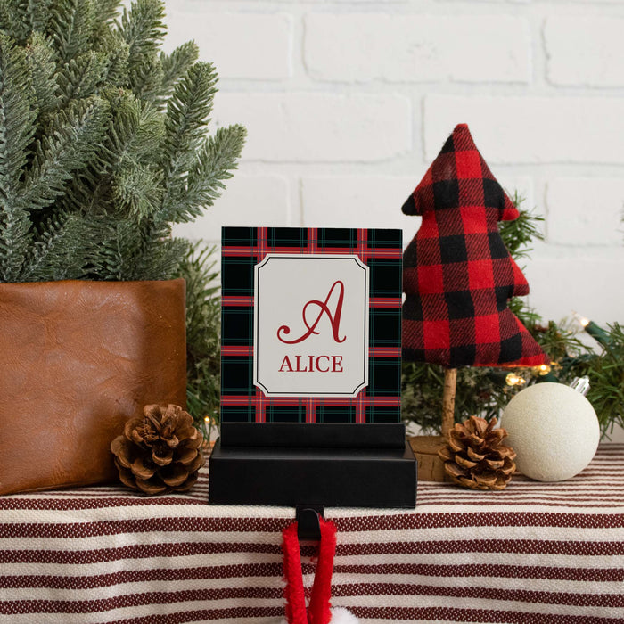 A personalized Christmas-themed stocking hanger featuring a black and red plaid background with a white central panel and the name 'ALICE' under a red script letter 'A'. The sign is styled on a striped table runner with pinecones, a red-brown leather bag, a red-and-black plaid Christmas tree decoration, and greenery against a white brick wall.