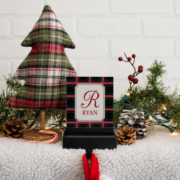 A personalized Christmas stocking hanger hook with a black and red plaid background, a white center panel, and the name 'RYAN' beneath a red monogram letter 'R'. The sign is styled on a white faux-fur surface with a small plaid Christmas tree, pinecones, greenery, and warm string lights against a white brick wall.