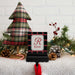 A personalized Christmas stocking hanger hook with a black and red plaid background, a white center panel, and the name 'RYAN' beneath a red monogram letter 'R'. The sign is styled on a white faux-fur surface with a small plaid Christmas tree, pinecones, greenery, and warm string lights against a white brick wall.
