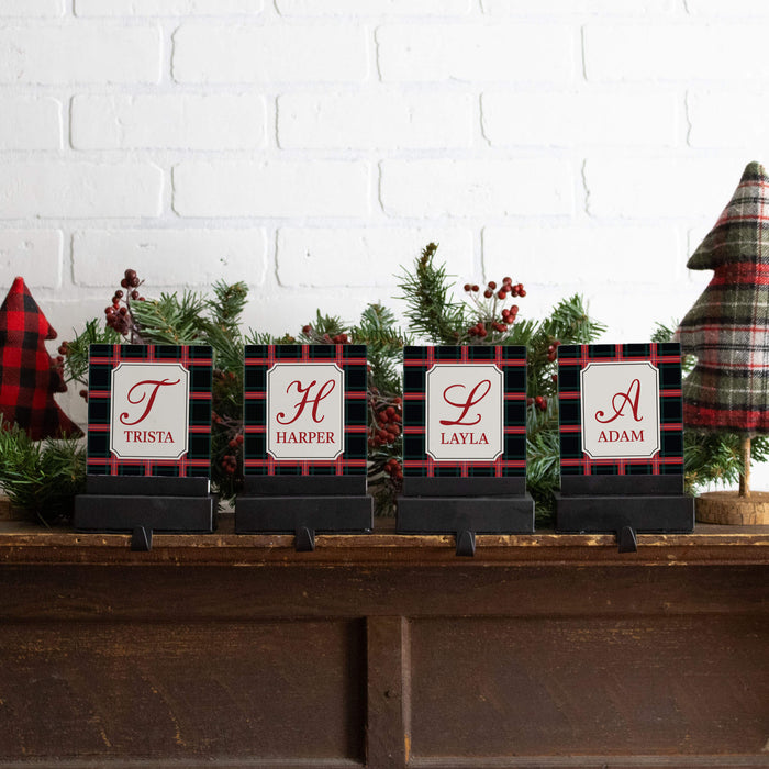 Four personalized black and red plaid stocking hanger name signs with white labels and red script monograms, displayed on a wooden mantel with holiday greenery and small trees against a white brick wall.