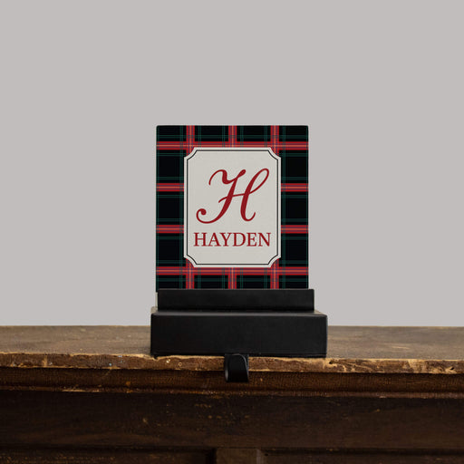 A personalized stocking hanger plaque with a black and red plaid pattern, featuring a cream central label with a red monogram 'H' and the name 'HAYDEN'. The sign is displayed on a black stand on a rustic wooden mantel against a solid gray background