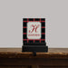 A personalized stocking hanger plaque with a black and red plaid pattern, featuring a cream central label with a red monogram 'H' and the name 'HAYDEN'. The sign is displayed on a black stand on a rustic wooden mantel against a solid gray background