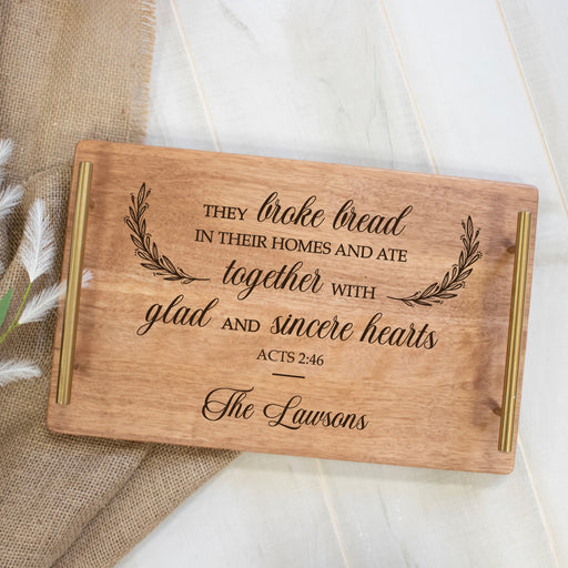 Wooden cutting board with engraved bible verse Acts 2:46 and gold accent handles on a neutral background of burlap and white wood