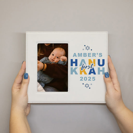 Hands holding a white picture frame featuring a photo of a baby and the name First Hannukah 2025