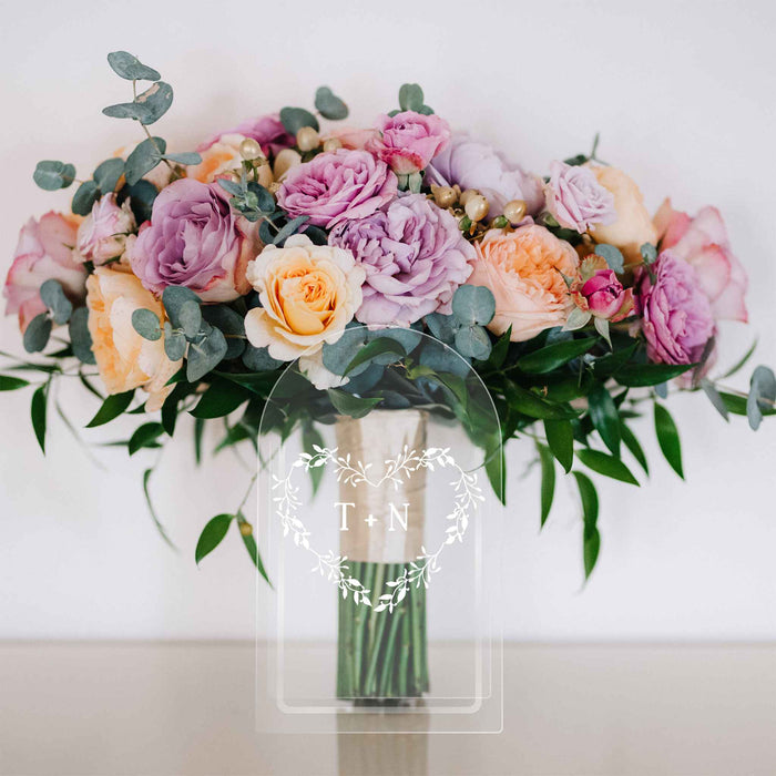 Clear acrylic vase with a monogrammed heart wreath holding colorful flowers