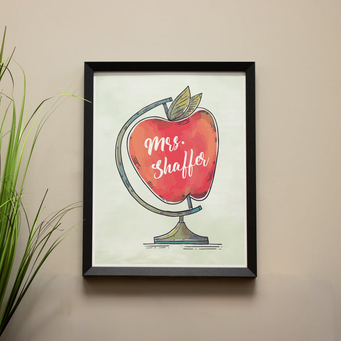 Framed artwork of a red apple with teacher's name on a beige wall.