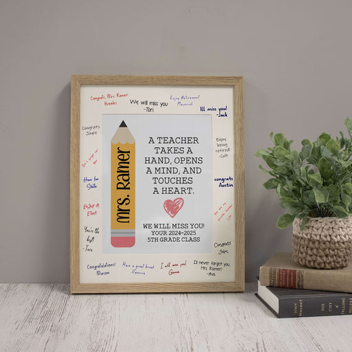 Framed print with a message for a teacher, placed on a surface with books and a plant.