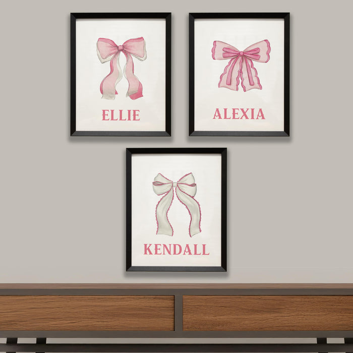 Three framed prints with pink bows and names on a wall above a wooden shelf.