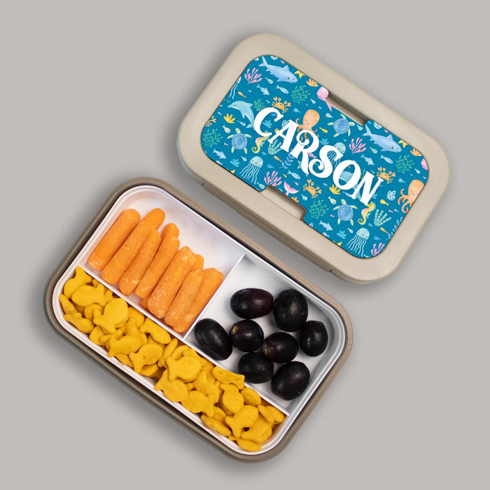 Personalized ocean-themed lunch box with compartments of snacks and a personalized lid on a gray background