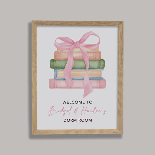 Framed artwork with books and a pink ribbon on a white background, displayed on a gray wall.