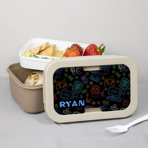 Lunch box with personalized gamer esign on the lid, containing snacks and a fork on a gray background