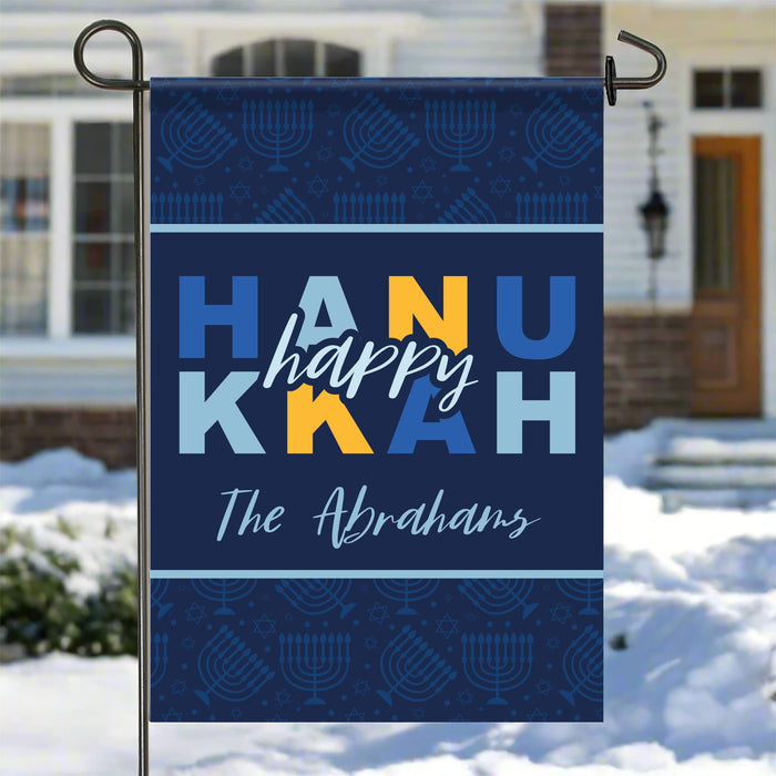 Personalized Happy Hanukkah garden flag with colorful text and menorah design on a blue background, displayed outdoors.