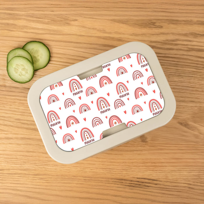 Personalized lunch box with a rainbow patterned lid on a wooden surface with cucumber slices.