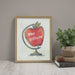 Framed artwork of a red apple with teacher's name on a wall next to books and a plant.