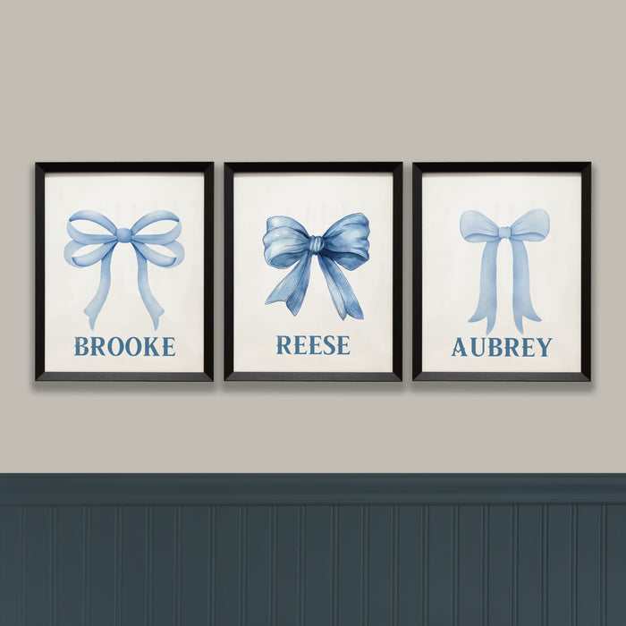 Three framed prints with blue bows and names on a gray wall.