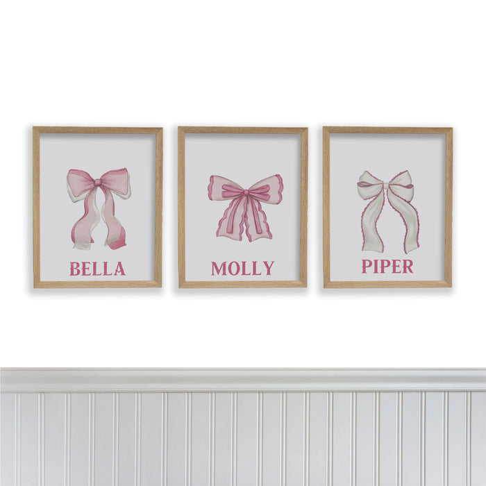 Three framed prints with pink bows and names on a white wall.