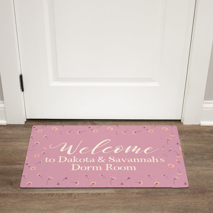 Pink dorm room doormat with floral designs and text on a wooden floor.