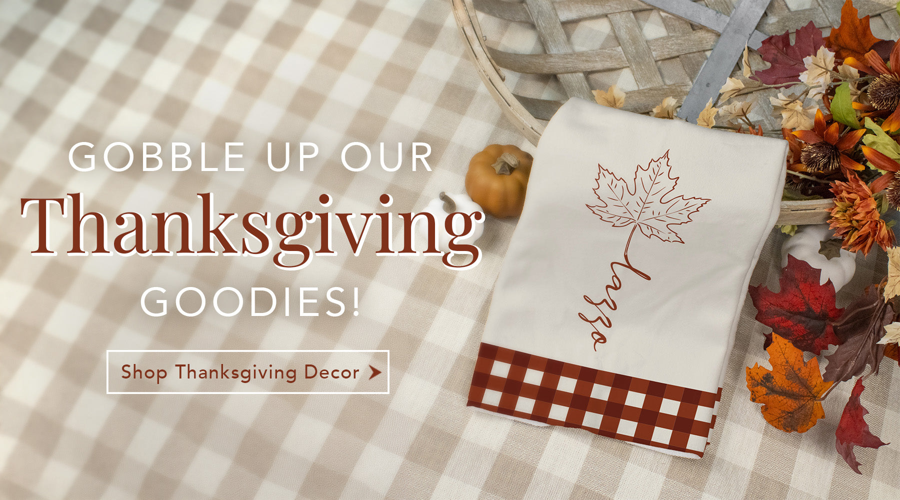 A white Thanksgiving-themed cloth napkin with a brown gingham check border and a brown maple leaf design, displayed on a beige-and-white gingham tablecloth alongside autumn leaves, a small pumpkin, and a basket. Large text on the left reads 'GOBBLE UP OUR Thanksgiving GOODIES! Shop Thanksgiving Decor'.