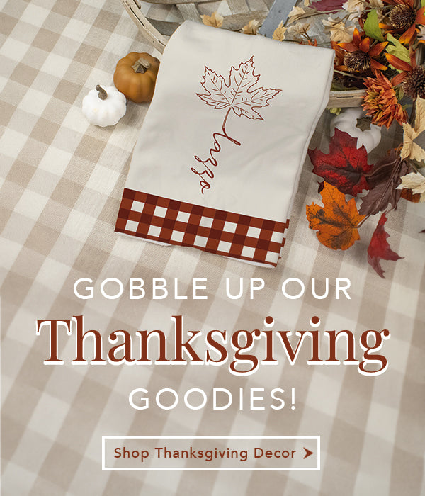 A white kitchen towel with a brown gingham band and maple leaf design, styled on a beige-and-white checkered tablecloth with pumpkins, autumn leaves, and a basket of fall decorations. Large text reads 'GOBBLE UP OUR Thanksgiving GOODIES!' with a smaller 'Shop Thanksgiving Decor' banner.