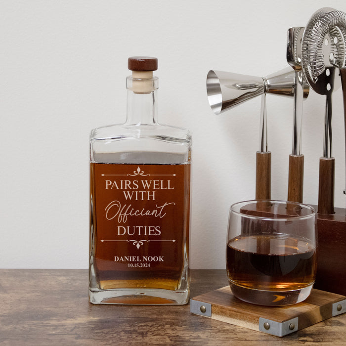 Personalized wedding officiant decanter next to a glass and cocktail shaker on a wooden surface.