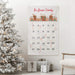 Custom family name advent calendar hanging on a wall next to a decorated Christmas tree and presents.