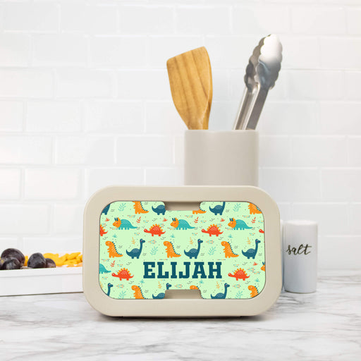 Children's lunch box with dinosaur pattern and name 'Elijah', next to a container with utensils on a marble surface.
