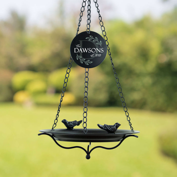 Black bird feeder with family last name branding against a blurred green garden background