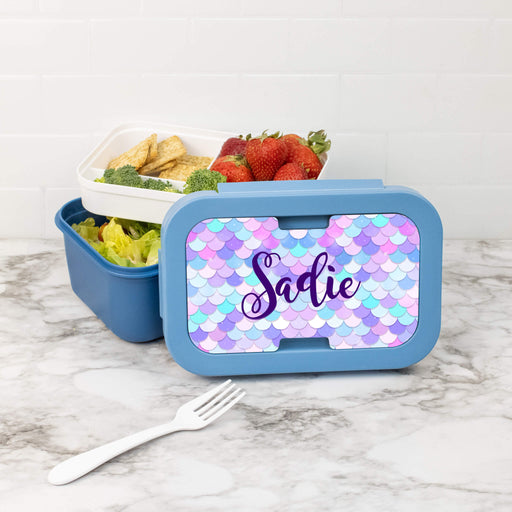 Blue lunch box with 'Sadie' on a mermaid scale pattern, containing snacks and a fork on a marble surface.