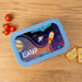 Space-themed lunchbox with 'REAGAN' printed on it, placed on a wooden surface with tomatoes and crackers.