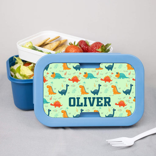 Personalized bento box with dinosaur pattern and 'OLIVER' text, containing a salad and fruit on a gray background.