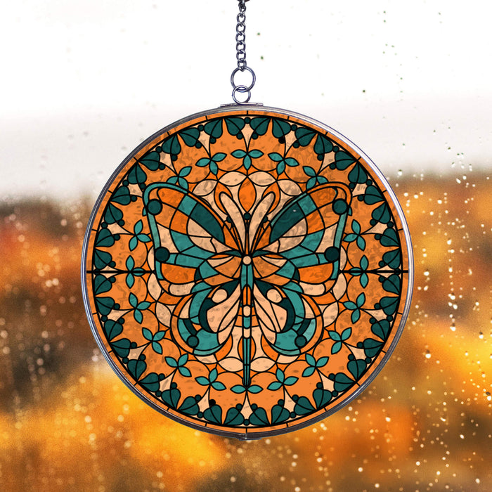 Decorative circular stained glass inspired acrylic suncatcher with orange and blue butterfly design, hanging against a rain covered window blurred background