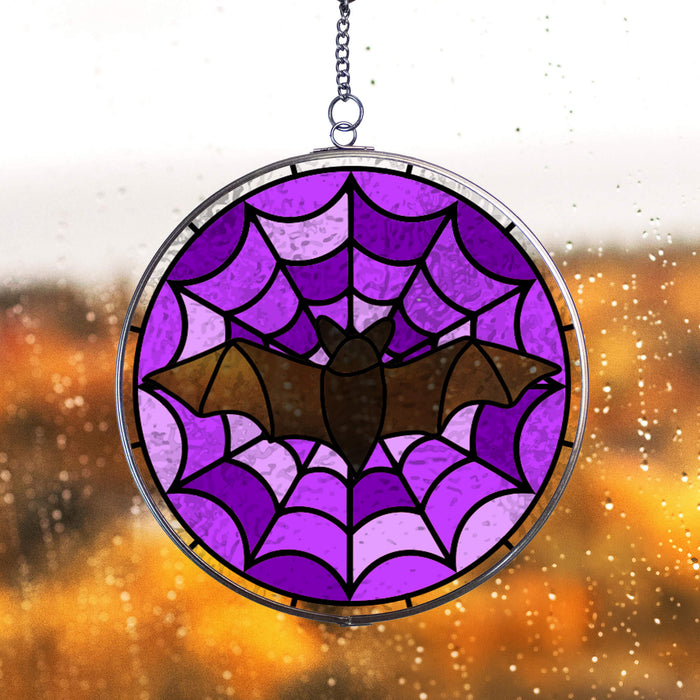 Stained glass window hanging light catcher with purple spider web and black bat design, against a blurred fall background.
